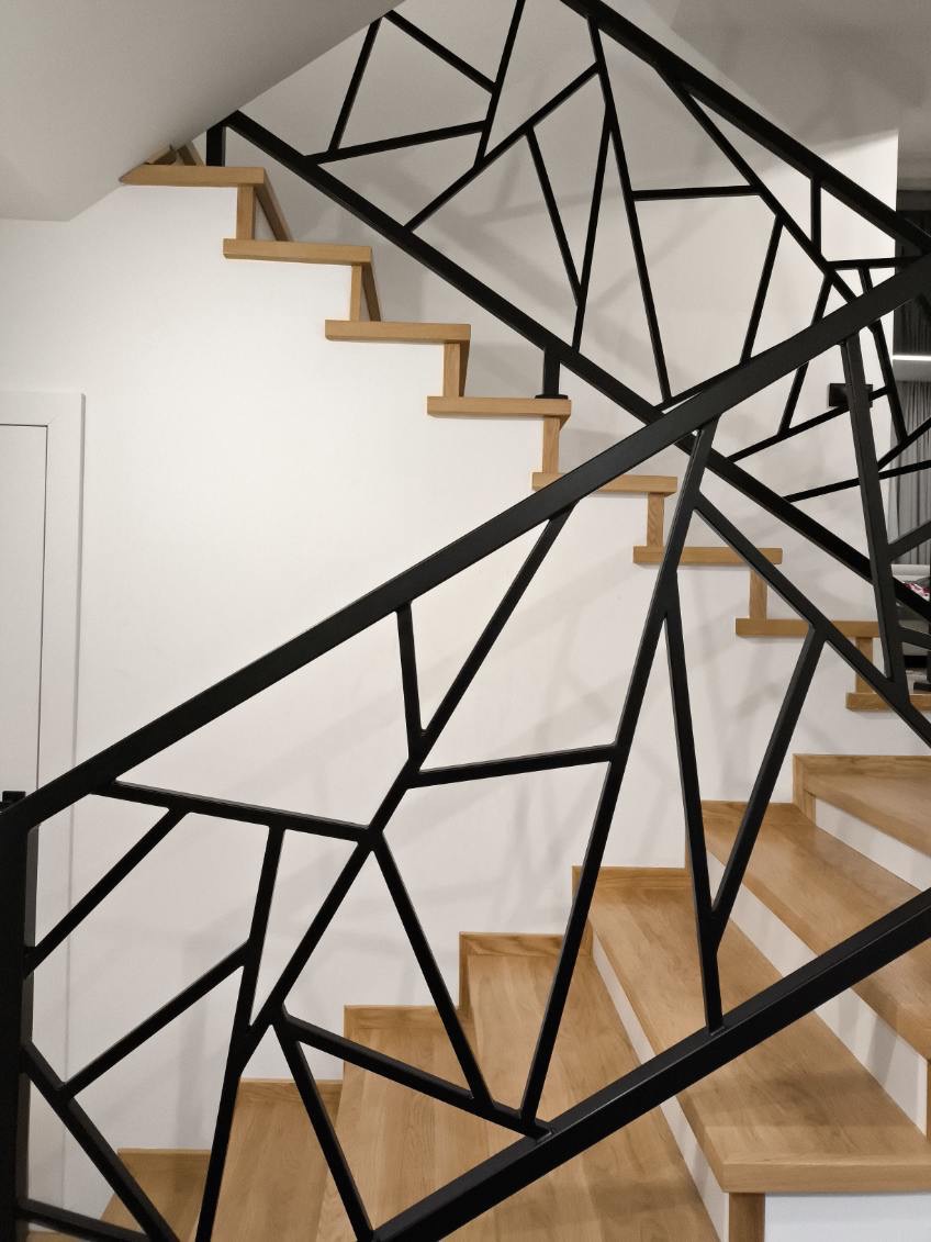 Nowoczesne schody z drewnianymi stopniami i metalową balustradą o geometrycznym wzorze w kolorze czarnym, widok od dołu.