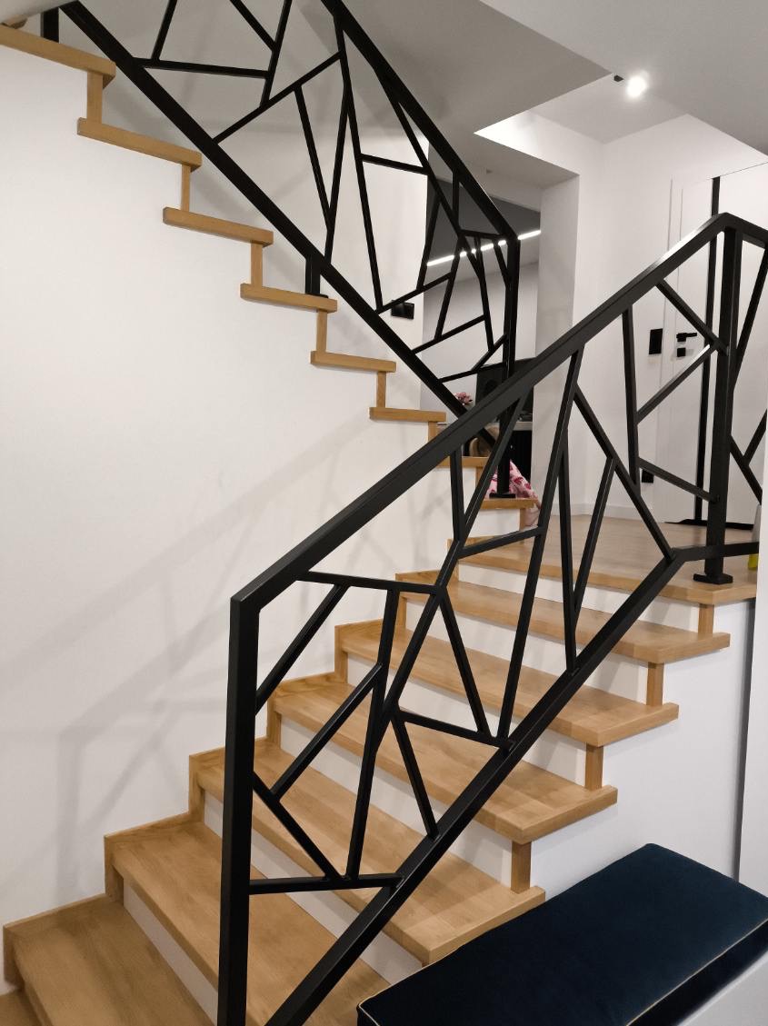 Schody drewniane z czarną, geometryczną balustradą metalową, widoczne z boku, z fragmentem niebieskiej pufy w dolnym rogu.