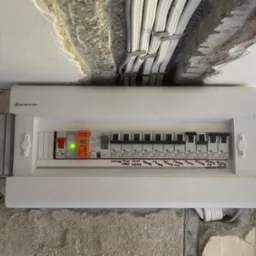 Rozdzielnia elektryczna marki Elektro-Plast z opisami obwodów (łazienka, salon, balkon) zamontowana na surowej ścianie. Widoczne kable w korycie.