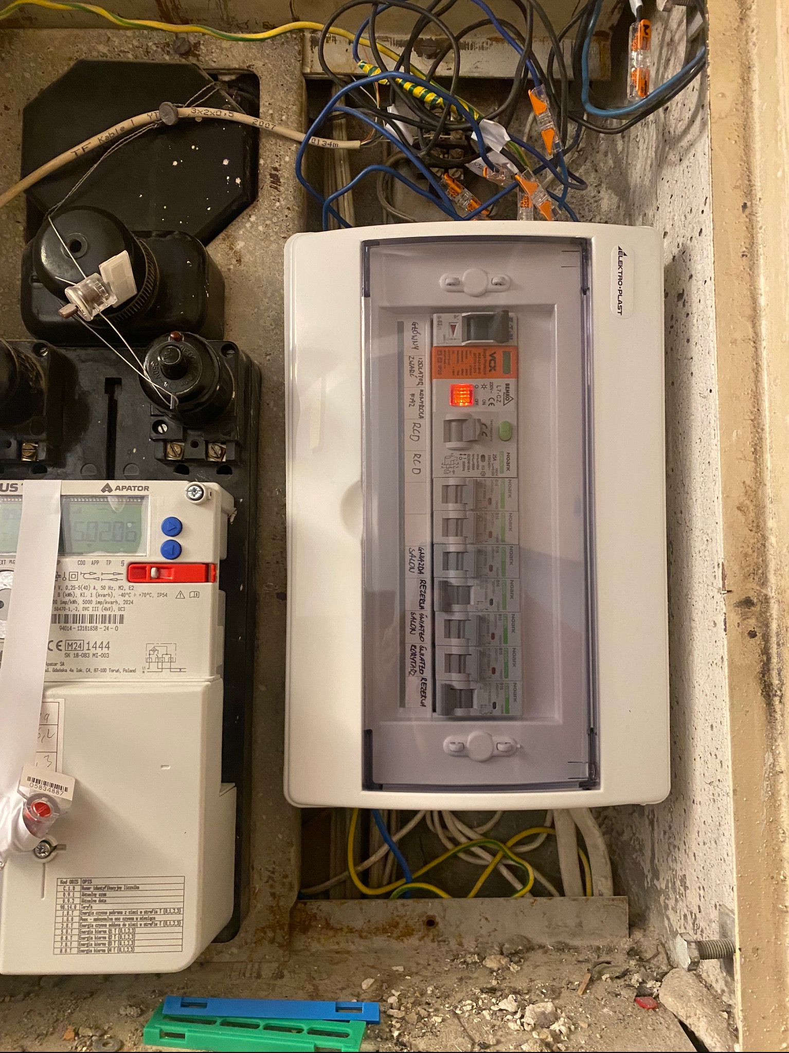 Otwarta skrzynka bezpiecznikowa Elektro-Plast z widocznymi bezpiecznikami NOARK, obok licznik energii elektrycznej APATOR, kable i przewody w ścianie.