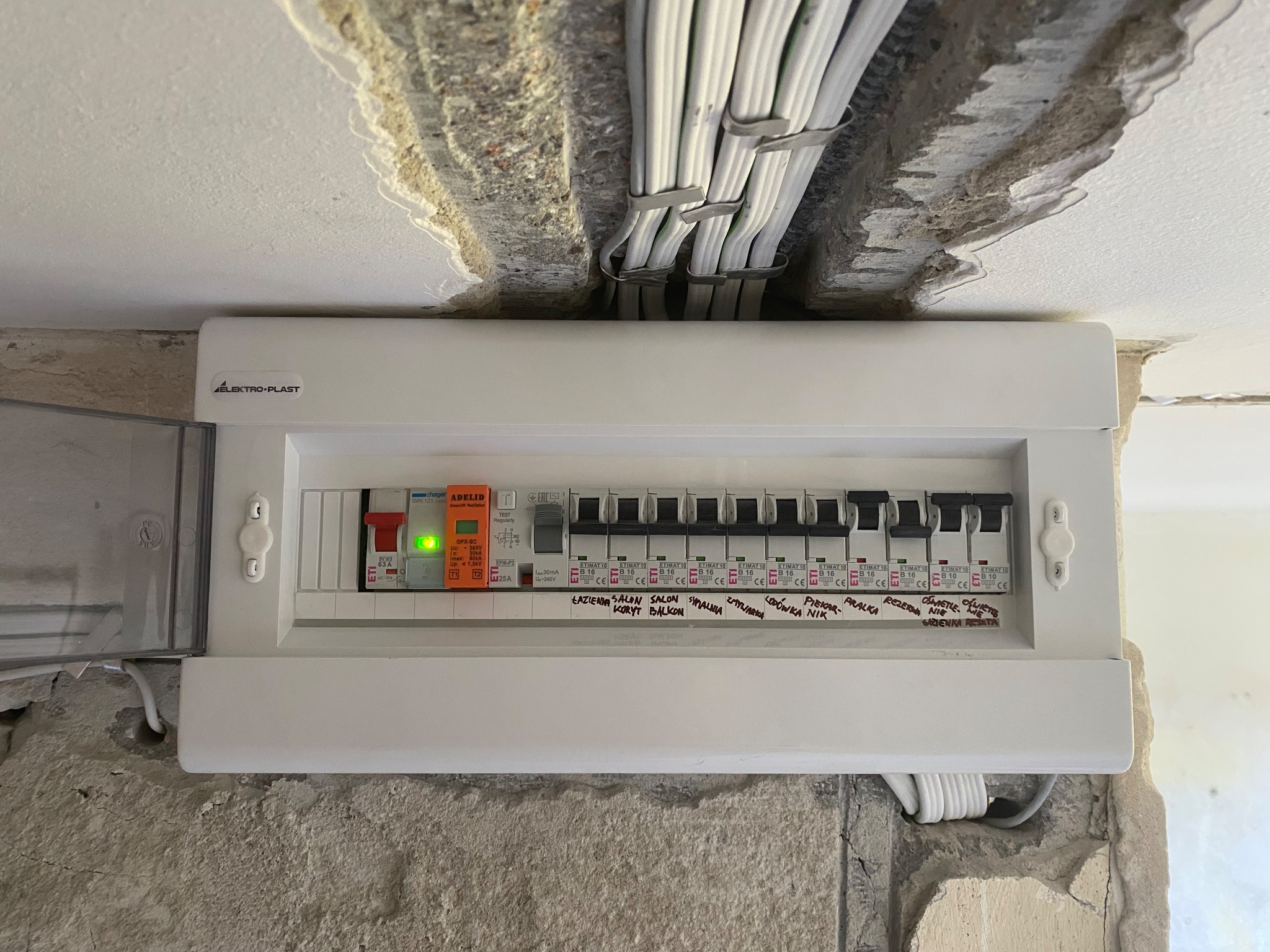 Rozdzielnia elektryczna marki Elektro-Plast z opisami obwodów (łazienka, salon, balkon) zamontowana na surowej ścianie. Widoczne kable w korycie.