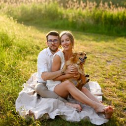 Karolina Klos Fotografia - Uśmiechnięta para siedzi na kocu na trawie w promieniach zachodzącego słońca, kobieta trzyma małego, brązowego psa.