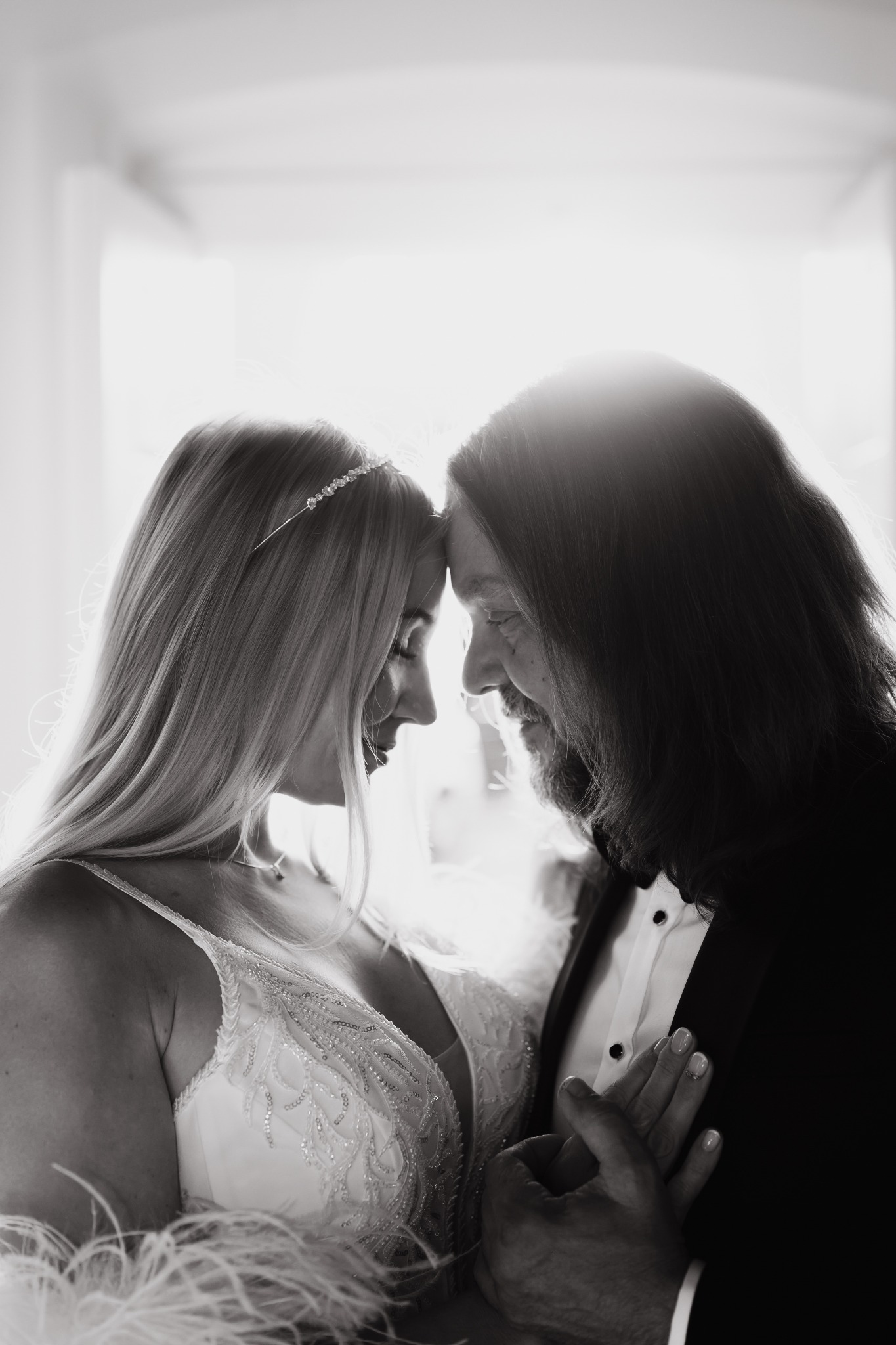 Czarno-białe zbliżenie na profil młodej pary, stykające się czołami, panna młoda w zdobionej sukni i tiarze, pan młody w smokingu z muszką, delikatne światło zza nich.