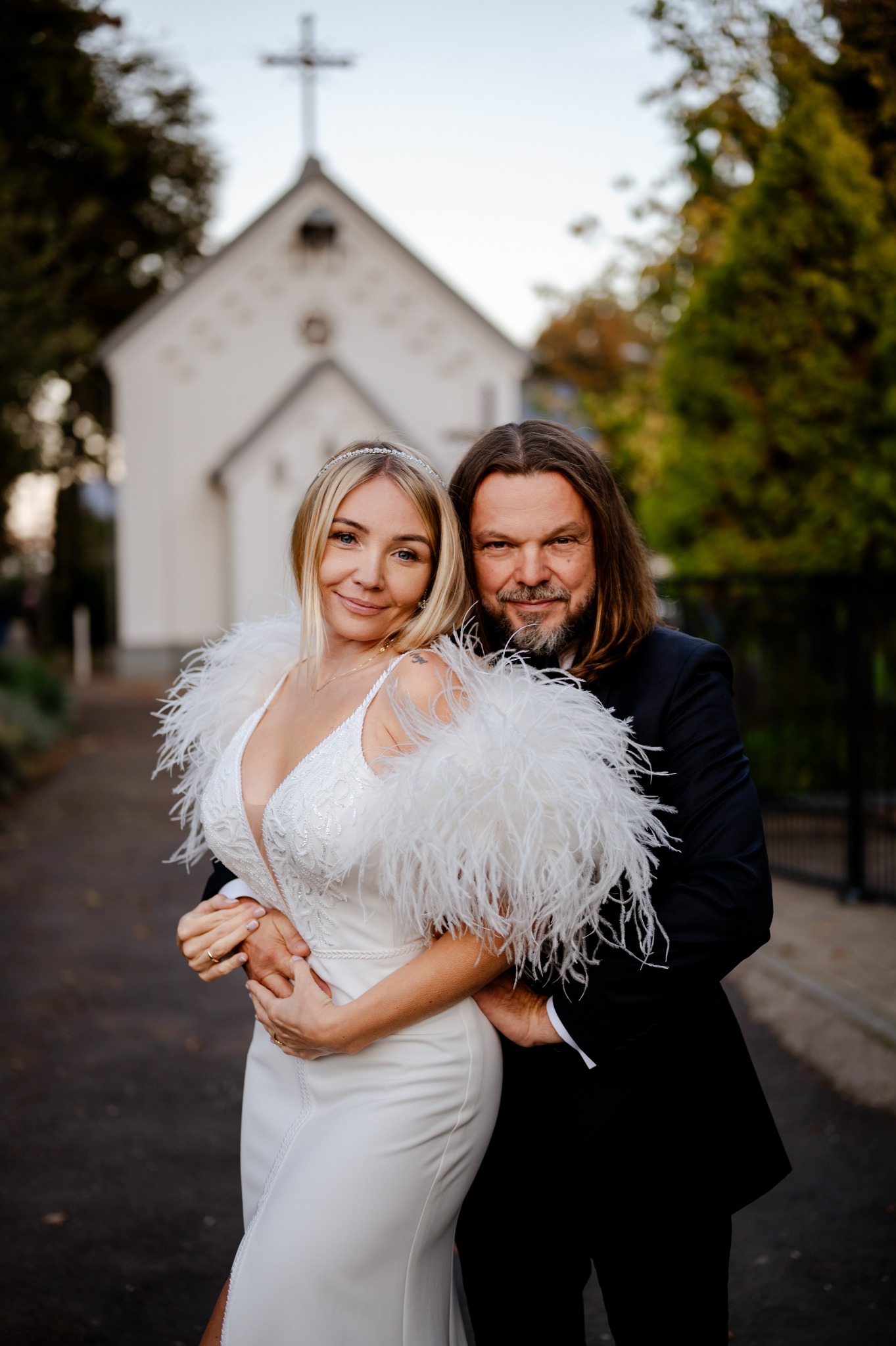 Para młoda w objęciach na tle białego kościoła z krzyżem; panna młoda w białej sukni z piórami i diademem, pan młody w czarnym garniturze.