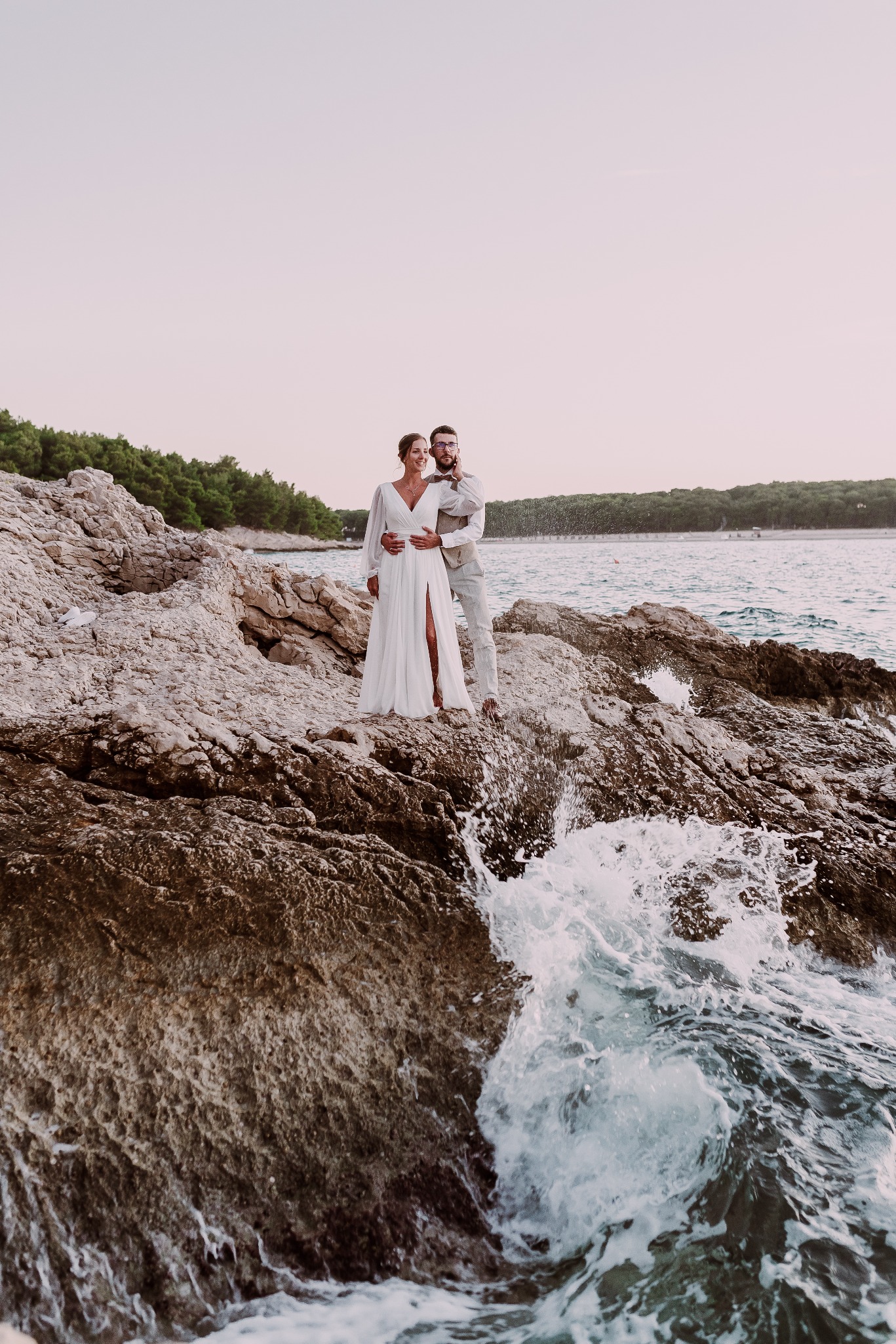Para młoda w objęciach na skalistym wybrzeżu, fale rozbijają się o skały, w tle zielony brzeg i spokojne morze, panna młoda w białej sukni z rozcięciem, pan młody w beżowej kamizelce i spodniach.
