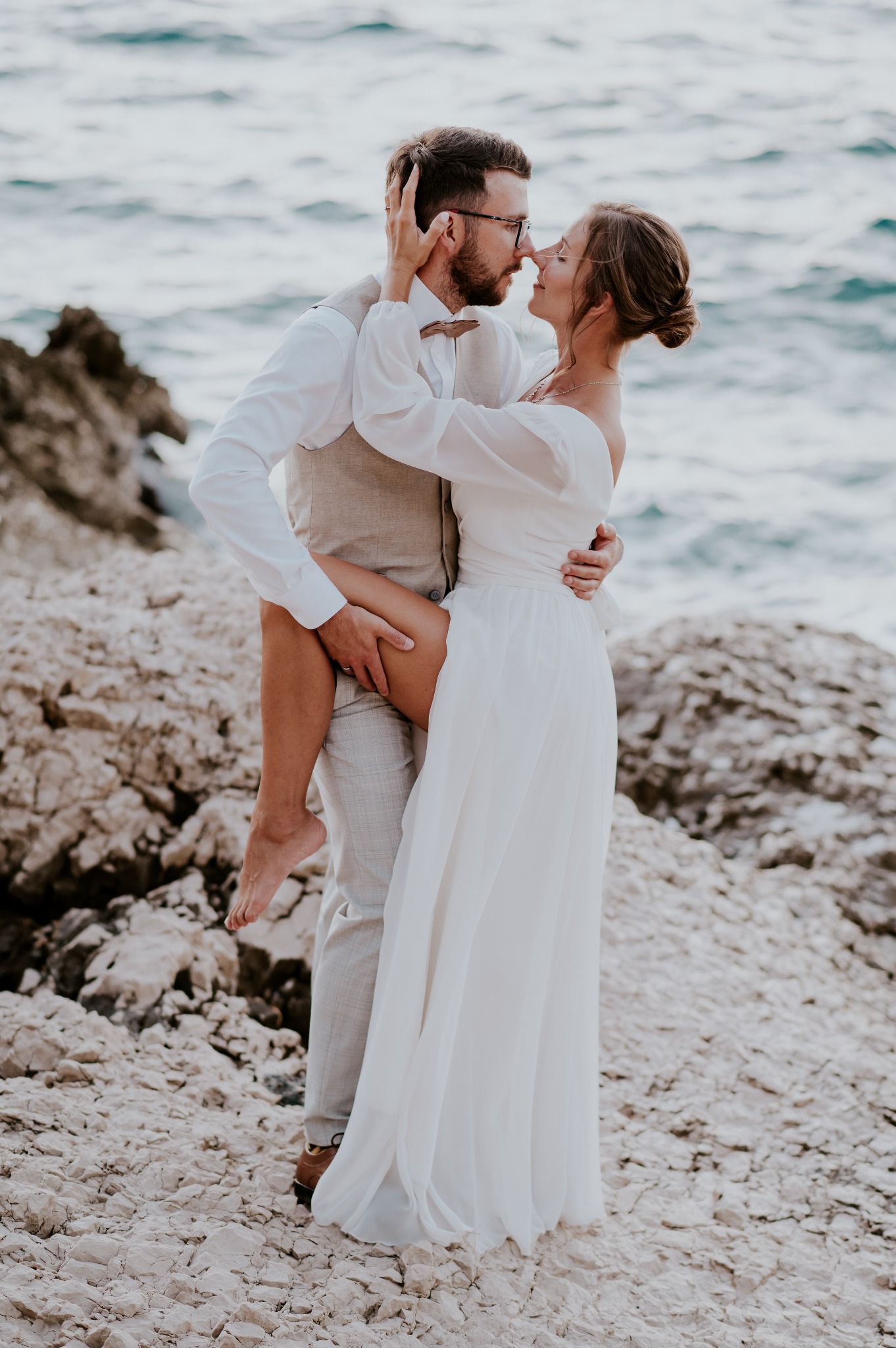 Para młoda w objęciach na skalistym wybrzeżu morza, panna młoda w zwiewnej białej sukni, pan młody w beżowej kamizelce i muszce, delikatne dotykanie nosami.