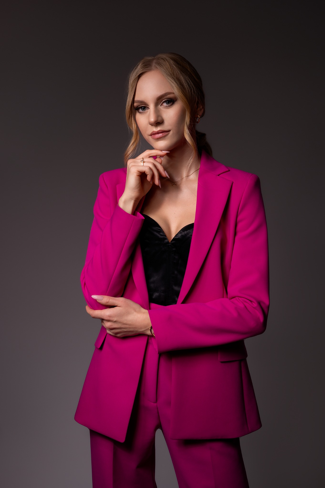 Portret kobiety w fuksjowym garniturze i czarnym gorsecie, z dłonią podpartą pod brodą, na szarym tle.