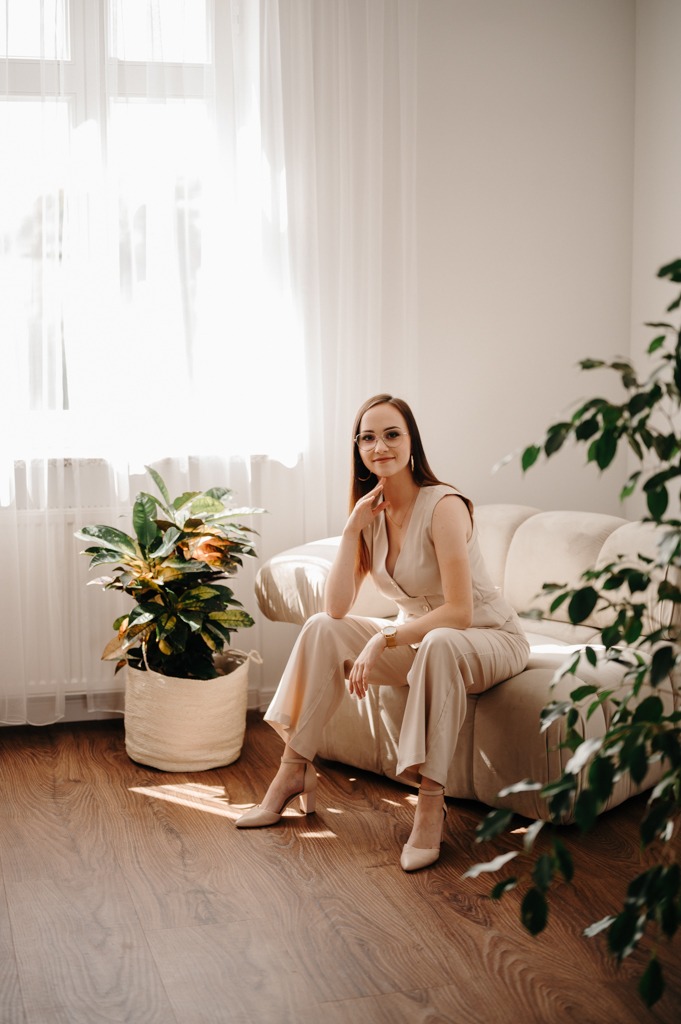 Kobieta w beżowym kombinezonie i okularach siedzi na kanapie w jasnym wnętrzu z roślinami doniczkowymi, pozując z ręką podpartą pod brodą.