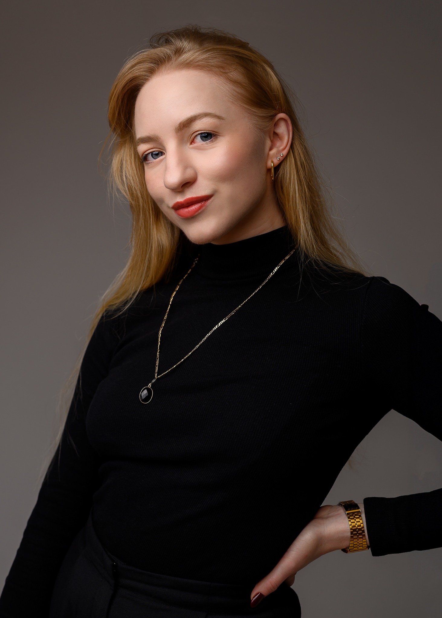Portret kobiety o jasnych włosach, ubrana w czarny golf i złotą biżuterię, pozująca na neutralnym tle, z delikatnym uśmiechem i subtelnym makijażem.