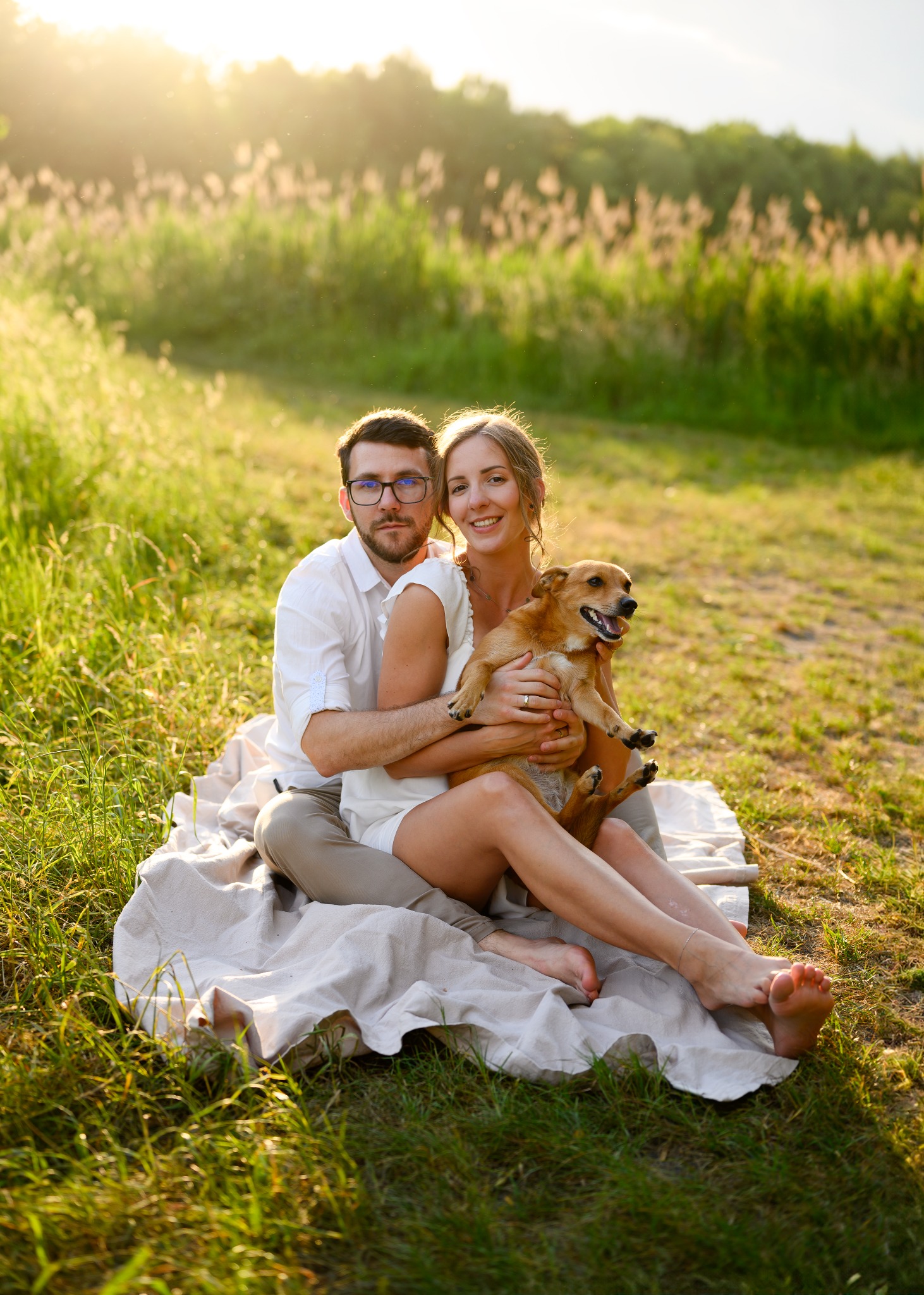 Uśmiechnięta para siedzi na kocu na trawie w promieniach zachodzącego słońca, kobieta trzyma małego, brązowego psa.