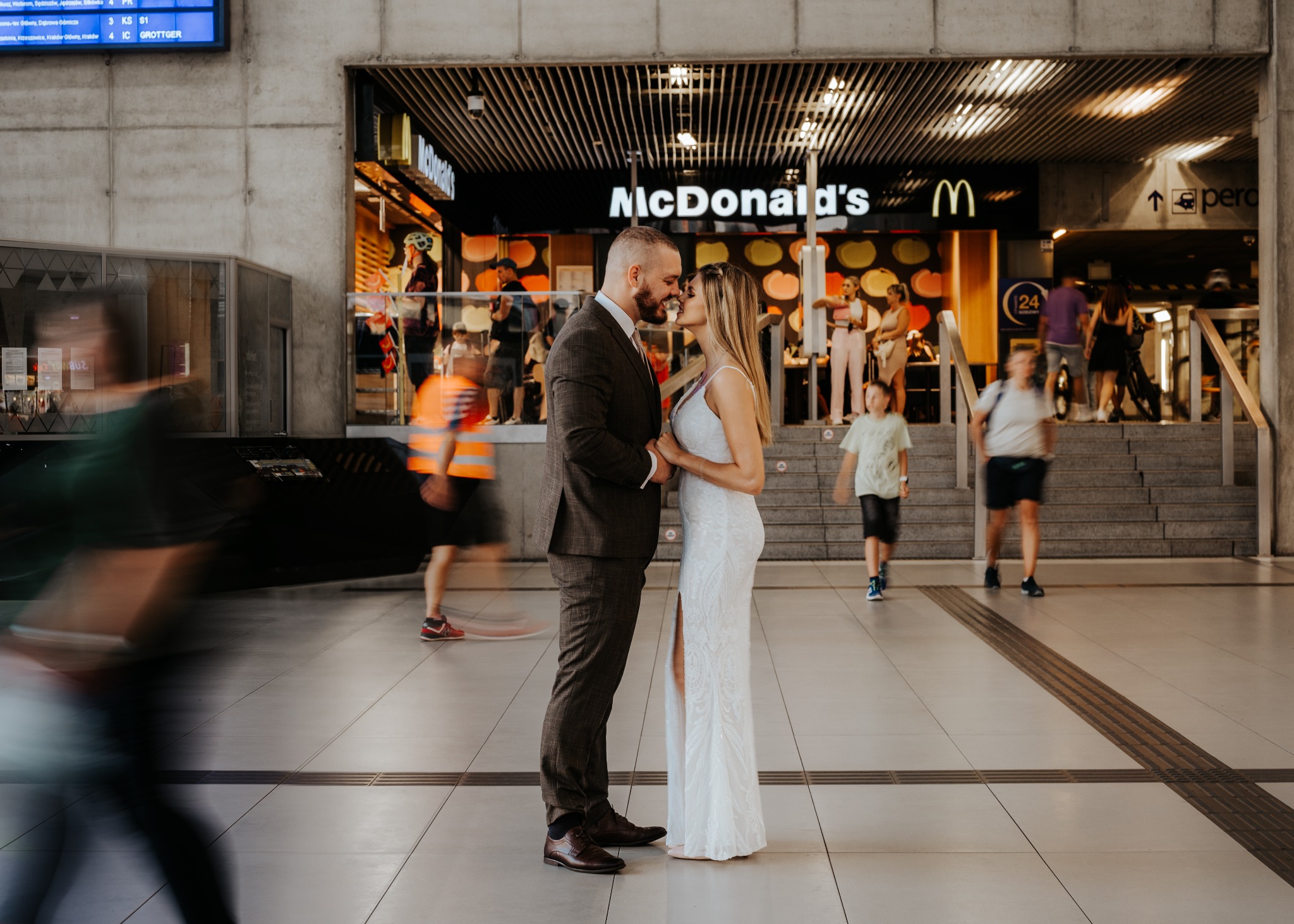 Para młoda w garniturze i sukni ślubnej, delikatnie całująca się na tle ruchliwej stacji kolejowej z widocznym McDonald's, tworząca kontrast między intymnością chwili a miejskim zgiełkiem.