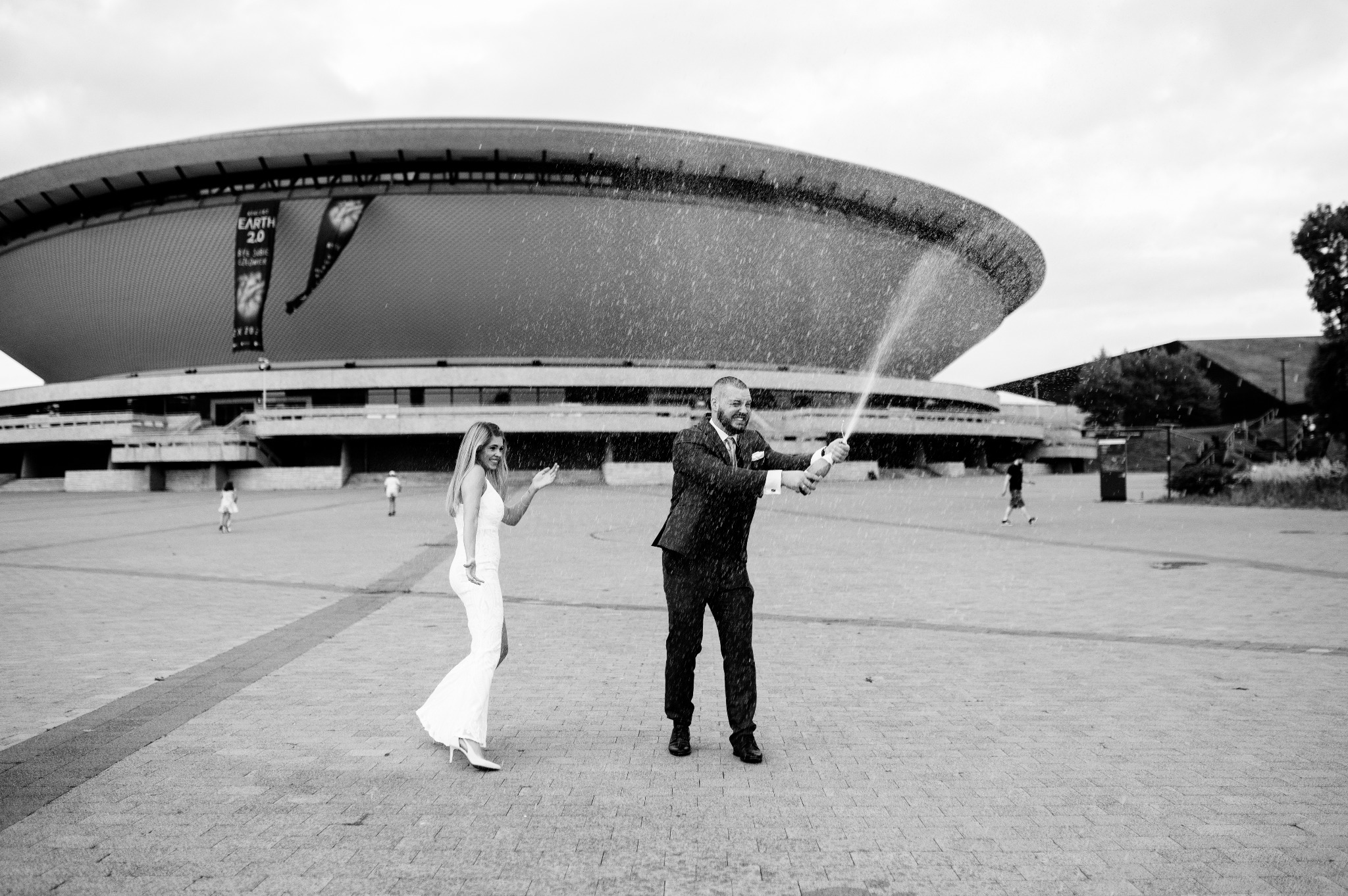 Czarno-białe zdjęcie pary świętującej z szampanem na tle Spodka w Katowicach. Panna młoda w długiej białej sukni, pan młody w garniturze, oboje uśmiechnięci.