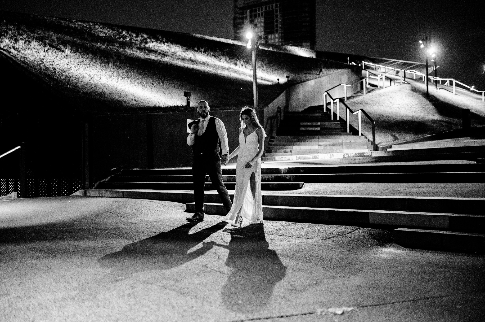 Czarno-białe zdjęcie pary młodej spacerującej po betonowym placu w nocy, mężczyzna trzyma marynarkę na ramieniu, a kobieta ma na sobie suknię z rozcięciem, w tle widoczne schody i oświetlenie...