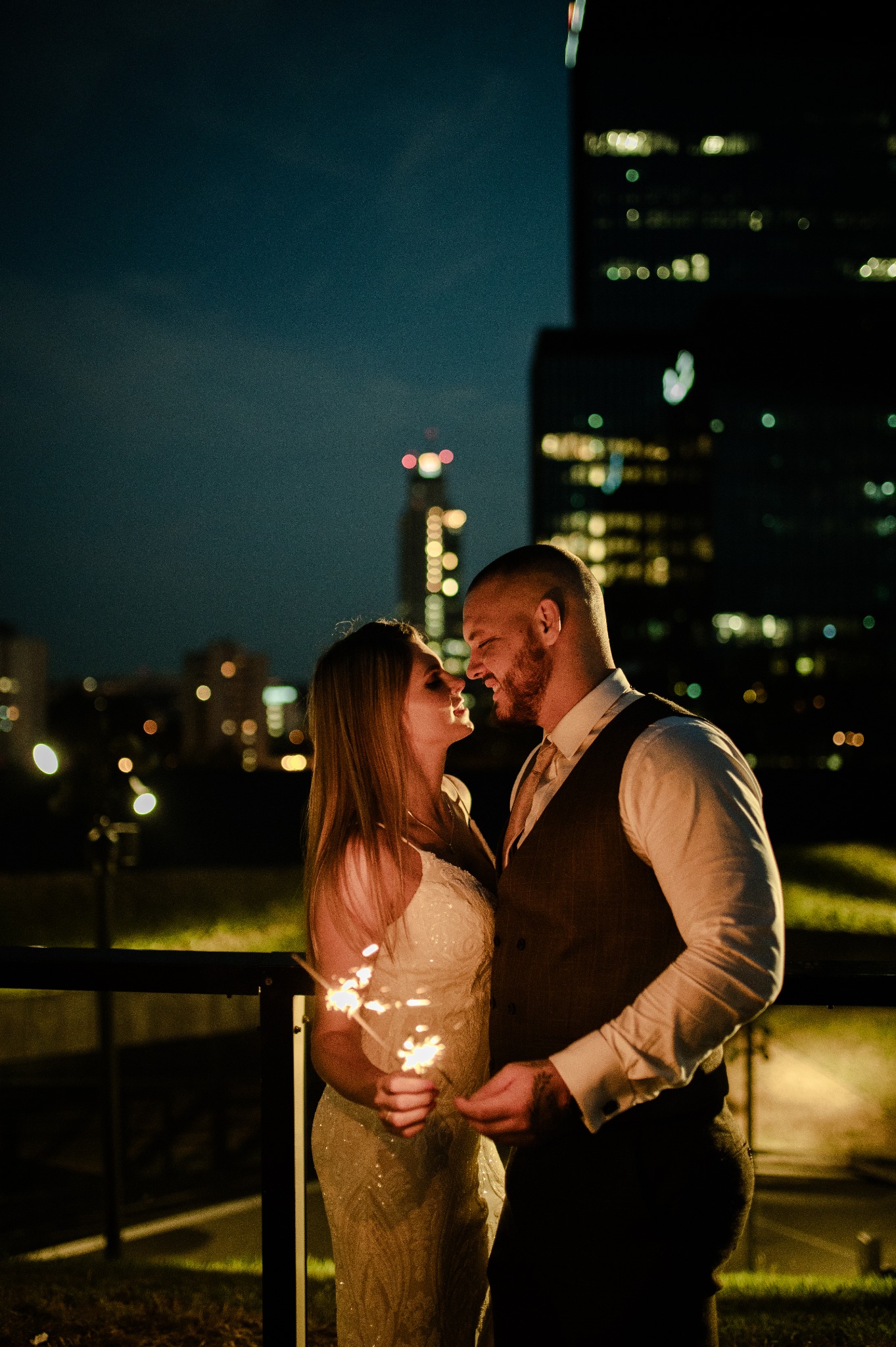 Para młoda, ona w sukni z cekinami trzyma zimne ognie, on w kamizelce, na tle nocnego miasta z wieżowcami.