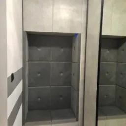 Wnęka z siedziskiem i tapicerowanymi panelami ściennymi w szarym odcieniu, z szafkami u góry i lustrem po prawej stronie. Nowoczesny design.