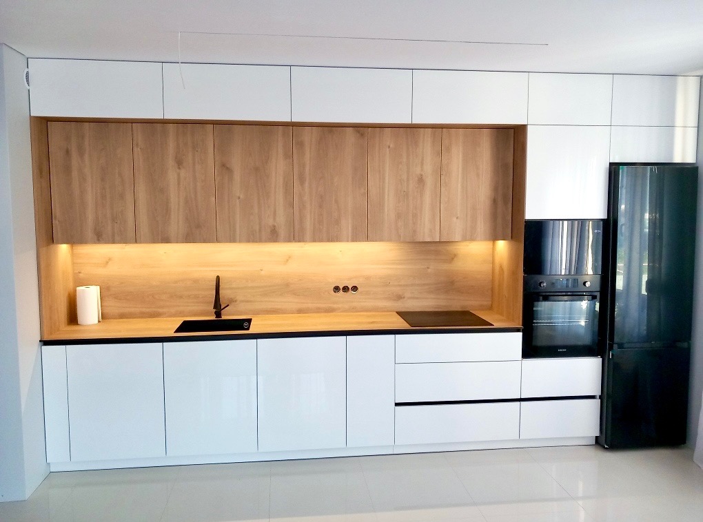 Nowoczesna kuchnia z białymi, lakierowanymi frontami dolnymi i drewnianymi górnymi. Oświetlenie LED pod szafkami. Czarny zlew i płyta indukcyjna. Czarna lodówka.