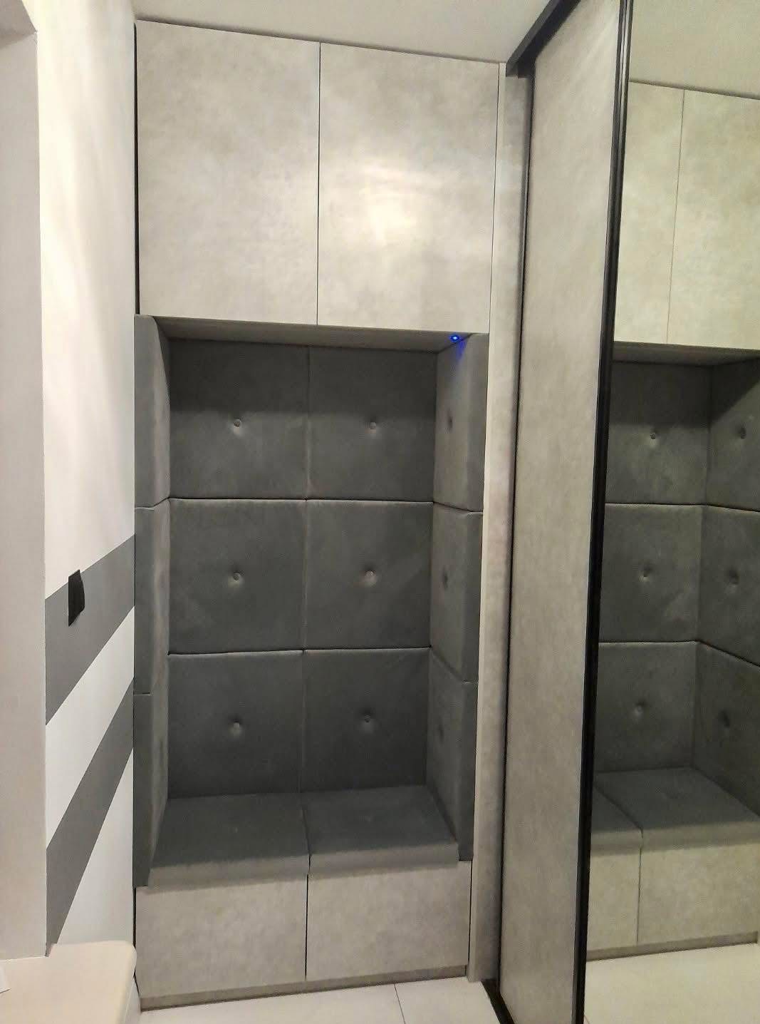 Wnęka z siedziskiem i tapicerowanymi panelami ściennymi w szarym odcieniu, z szafkami u góry i lustrem po prawej stronie. Nowoczesny design.