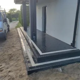 Nowoczesne schody wejściowe z czarnej kostki brukowej, wykończone białymi pasami, prowadzące do jasnego budynku. Widoczny fragment samochodu dostawczego i teren przed domem.