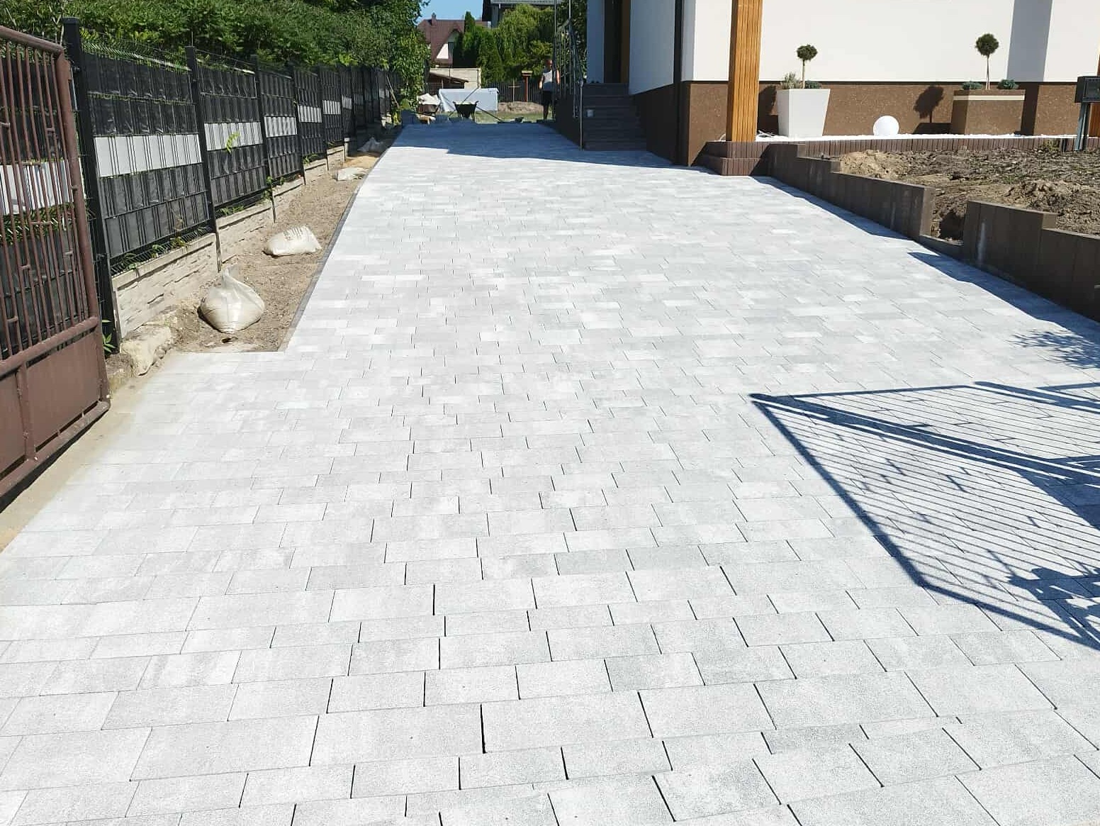 Jasnoszara kostka brukowa ułożona na podjeździe, widok na dom z elementami drewnianymi i czarnym ogrodzeniem. W tle widać wózek budowlany.