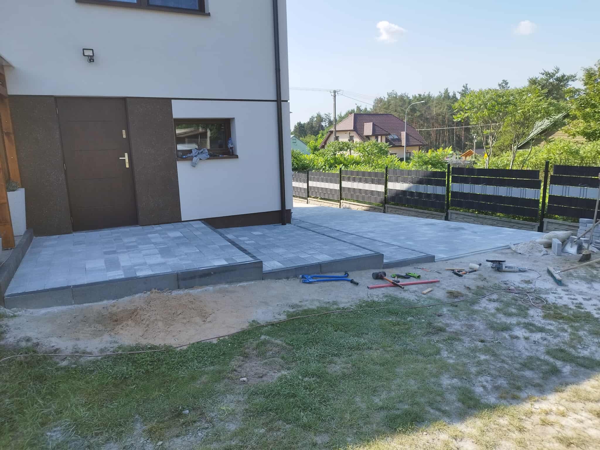 Ułożona szara kostka brukowa przed nowoczesnym domem. Widoczne narzędzia brukarskie i fragment trawnika. Kostka tworzy podjazd i chodnik.