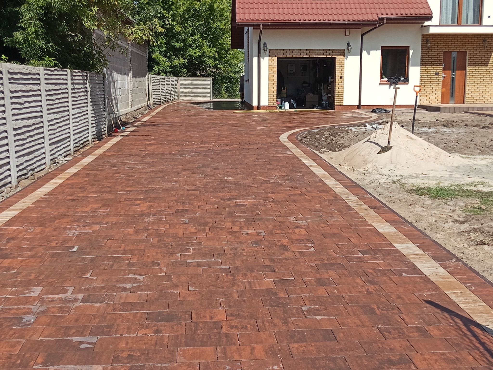 Ceglana kostka brukowa z jasnymi obrzeżami tworzy ścieżkę do garażu. Na piasku leżą narzędzia: łopata i szpadel. W tle dom z czerwoną dachówką i betonowy płot.