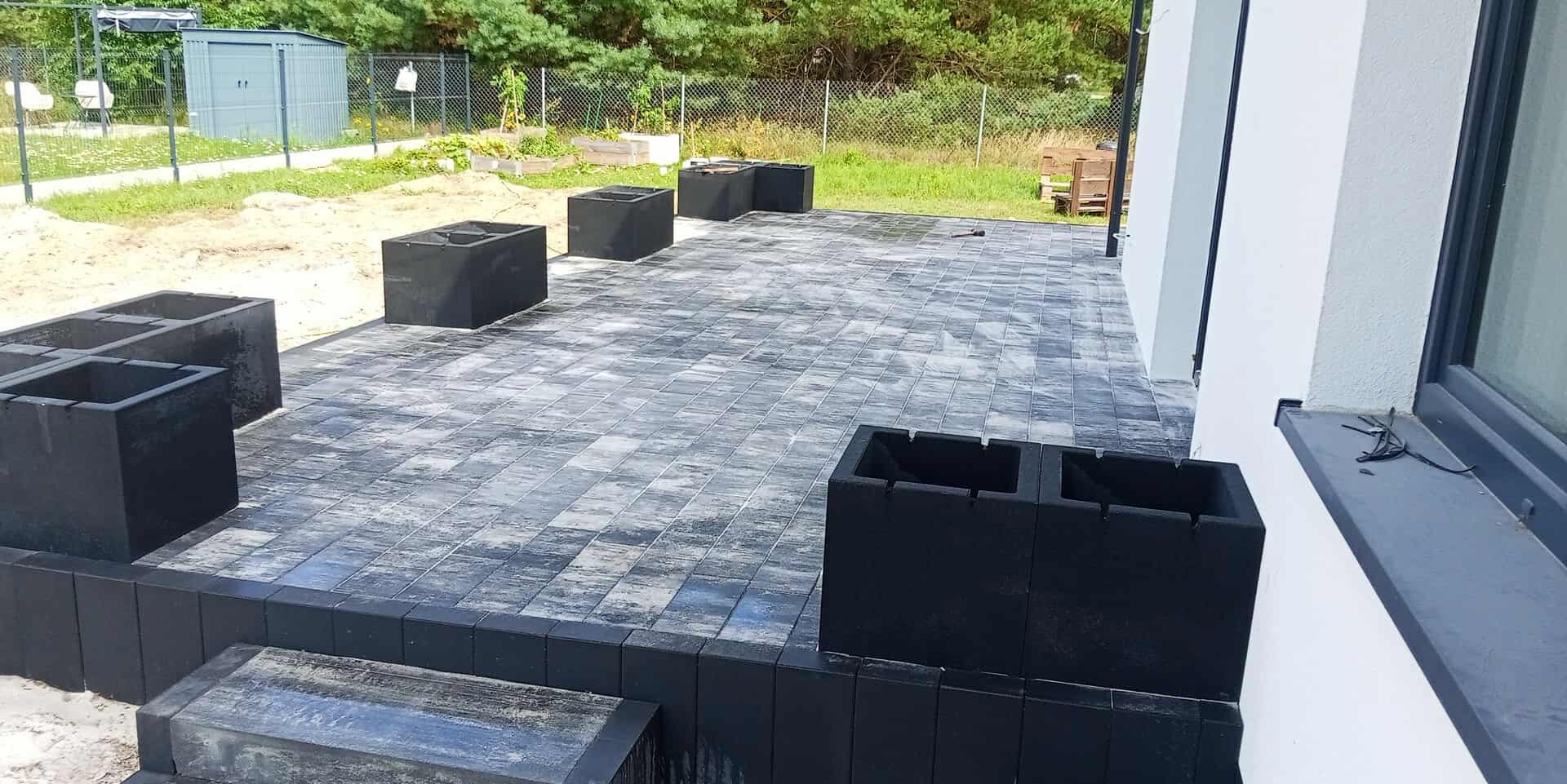 Ułożona szara kostka brukowa na tarasie z czarnymi donicami, widok na ogród z domkiem narzędziowym w tle. Nowoczesne wykończenie przestrzeni.