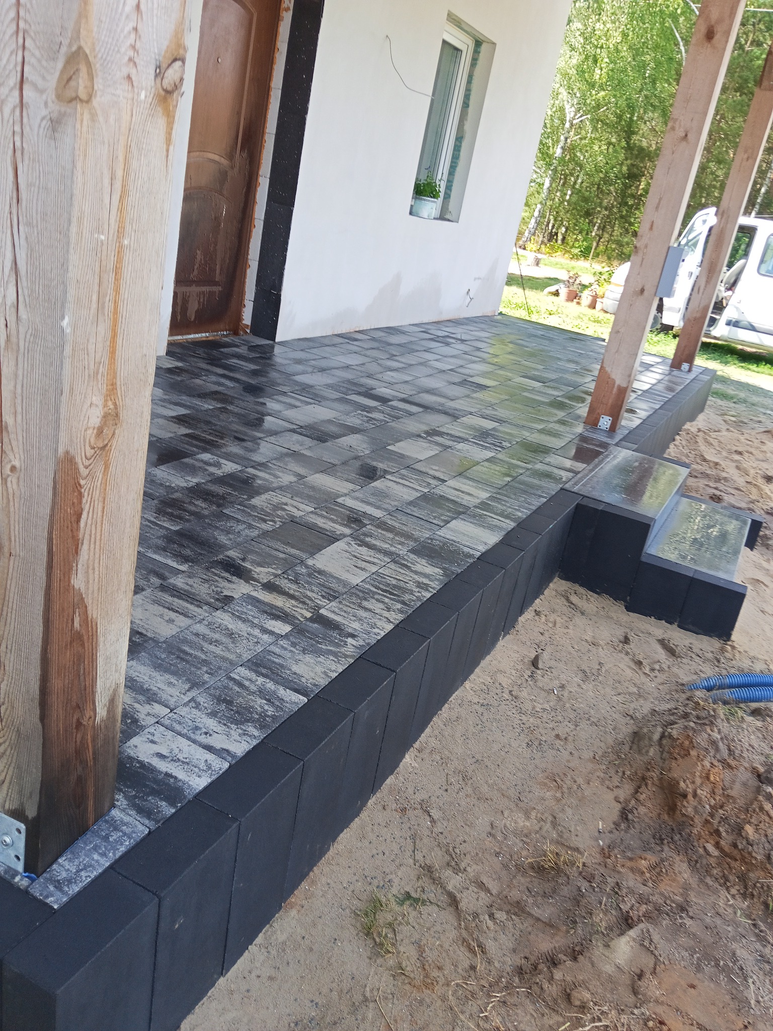 Ułożona kostka brukowa w odcieniach szarości z czarnym obrzeżem, schody z kostki, widok na dom w Siedlcach. Elementy drewniane konstrukcji zadaszenia.