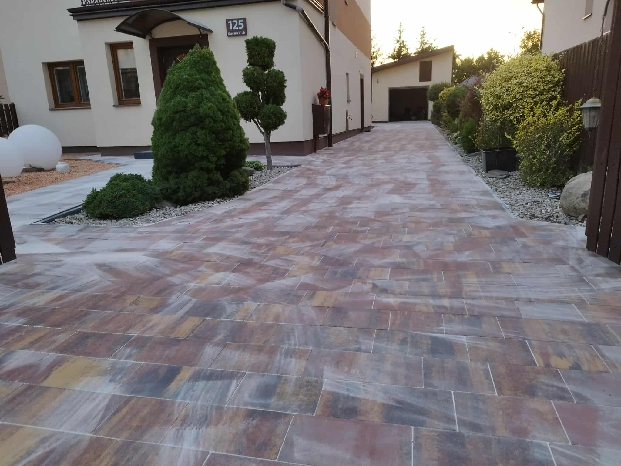 Kolorowa kostka brukowa na podjeździe domu z numerem 125, z zadbaną zielenią i garażem w tle, prezentuje estetyczne wykończenie posesji.