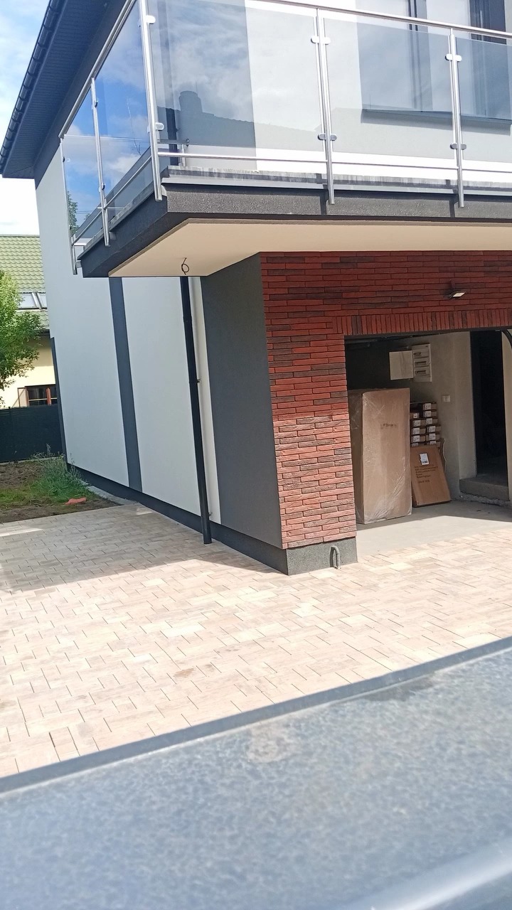 Nowoczesny dom z balkonem i kostką brukową. Elewacja w odcieniach bieli i szarości, z akcentami cegły. Widoczny fragment posesji i garaż z zapakowanymi elementami.