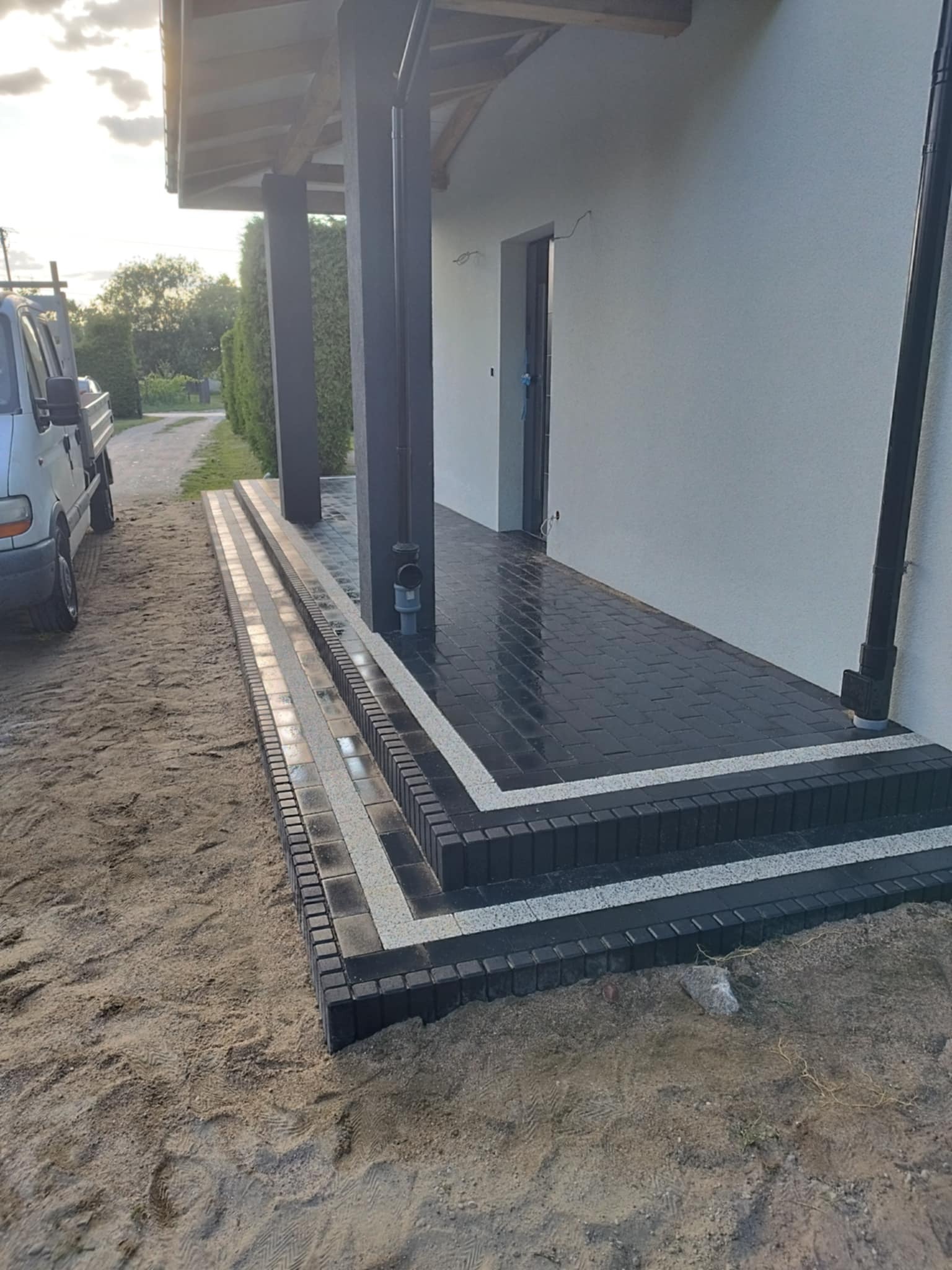Nowoczesne schody wejściowe z czarnej kostki brukowej, wykończone białymi pasami, prowadzące do jasnego budynku. Widoczny fragment samochodu dostawczego i teren przed domem.