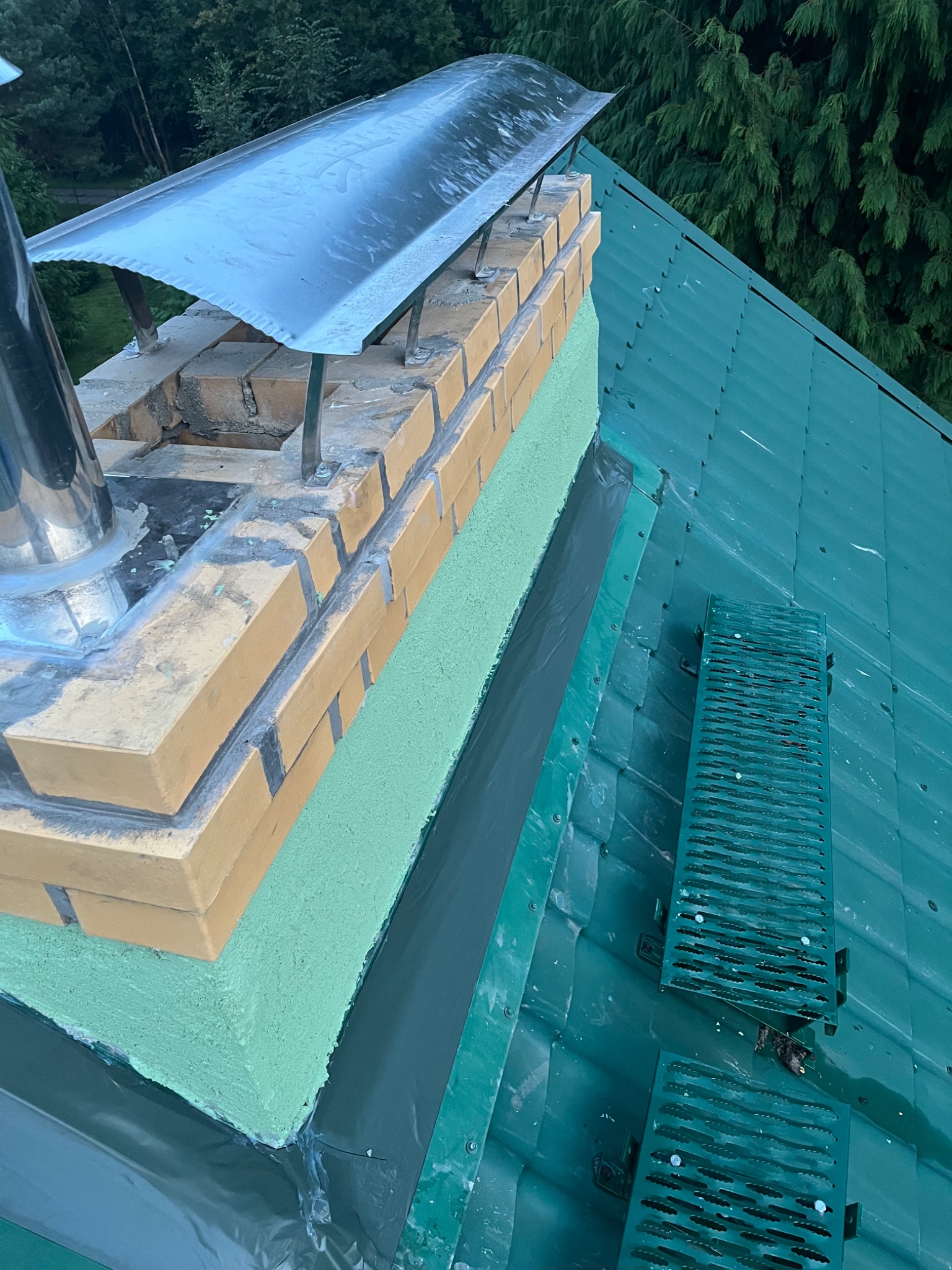 Kompleksowy remont komina z cegły klinkierowej, ocieplonego pianką, z metalowym daszkiem i nową obróbką blacharską na zielonym dachu z drabinkami.