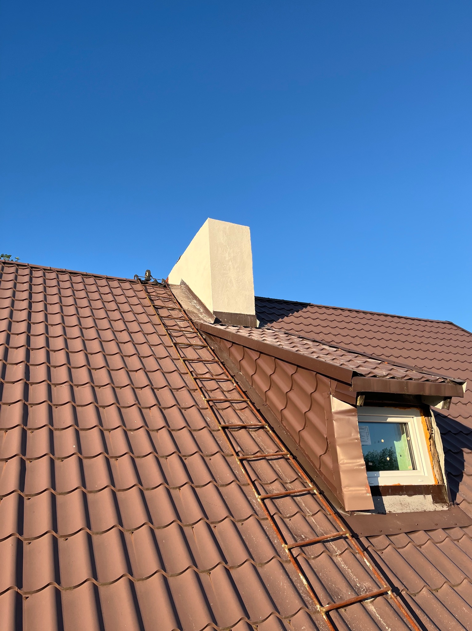 Brązowy dach z dachówki ceramicznej z oknem dachowym i kominem, drabina oparta o dach, błękitne niebo w tle. Widok z dołu.