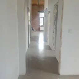 Długi korytarz w trakcie remontu z wylewką samopoziomującą, widoczne ślady po szalunkach na ścianach i surowa cegła w tle, światło wpadające przez okno.