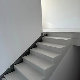 MOWA GROUP SPÓŁKA Z OGRANICZONĄ ODPOWIEDZIALNOŚCIĄ - Schody wewnętrzne pokryte jasnoszarą posadzką żywiczną, widoczne niedoskonałości wykończenia przy ścianie, ślady spływającej farby na ścianie bocznej.