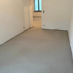 MOWA GROUP SPÓŁKA Z OGRANICZONĄ ODPOWIEDZIALNOŚCIĄ - Jasnoszara, gładka posadzka w nowym pomieszczeniu z białymi ścianami. Widoczne otwory na gniazdka. Okno w tle zasłonięte folią.