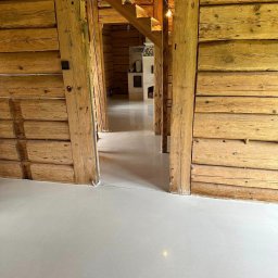 MOWA GROUP SPÓŁKA Z OGRANICZONĄ ODPOWIEDZIALNOŚCIĄ - Jasnoszara, gładka posadzka żywiczna w drewnianym domu z bali. Widok na wnętrze przez drewniane framugi, schody i kominek w tle. Nowoczesne wykończenie w rustykalnym otoczeniu.