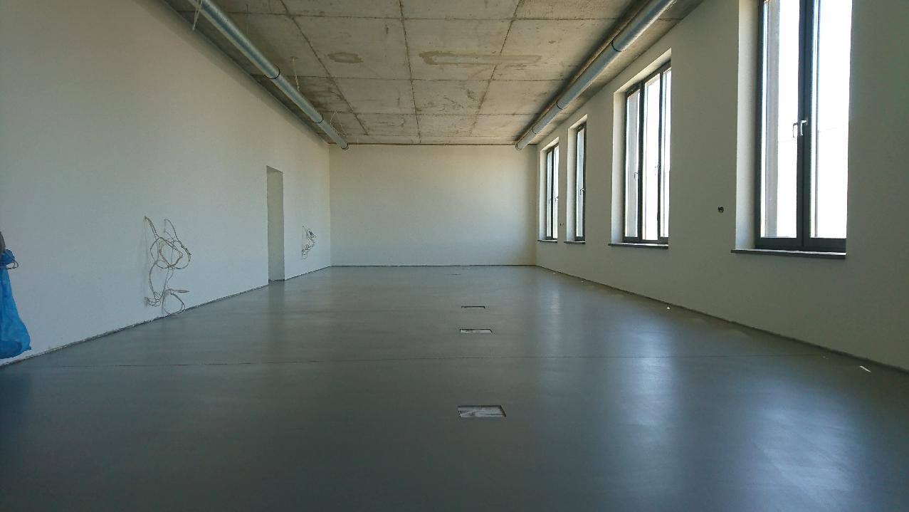 Szara, gładka posadzka w surowym, industrialnym wnętrzu z betonowym stropem i rurami pod sufitem. Widoczne okna z naturalnym światłem i wystające przewody ze ściany.