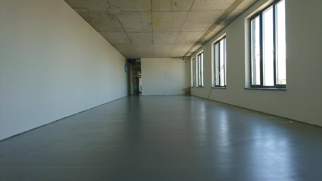 Wnętrze pustego pomieszczenia z szarą, błyszczącą posadzką, surowym betonowym sufitem i rzędem okien w czarnych ramach wzdłuż prawej ściany.