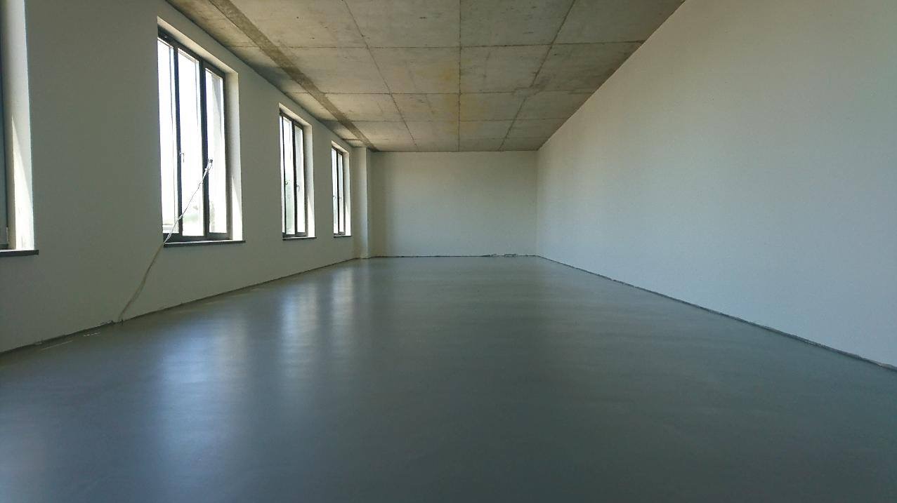 Szara, gładka posadzka w pustym pomieszczeniu z betonowym sufitem i rzędem okien po lewej stronie, odbijająca światło.