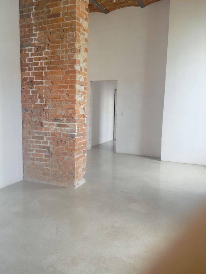 Jasnoszara, gładka posadzka w minimalistycznym wnętrzu z fragmentem ceglanego filara. Widoczne przejście do kolejnego pomieszczenia w tle.