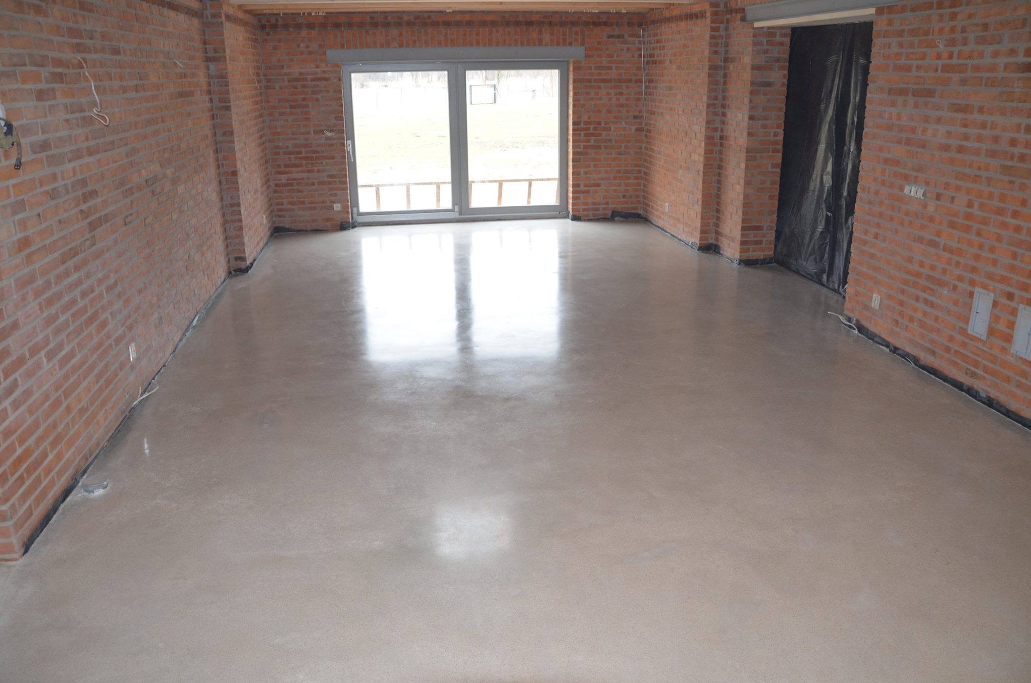 Szlifowana, jasnoszara posadzka w surowym wnętrzu z ceglanymi ścianami i dużym oknem balkonowym.