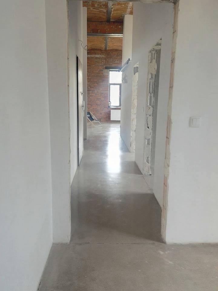 Długi korytarz w trakcie remontu z wylewką samopoziomującą, widoczne ślady po szalunkach na ścianach i surowa cegła w tle, światło wpadające przez okno.