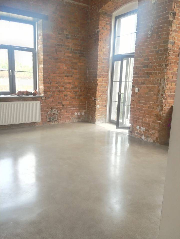 Polerowana, jasnoszara posadzka w surowym wnętrzu z ceglanymi ścianami i dużymi oknami. Minimalistyczny grzejnik pod oknem po lewej stronie.