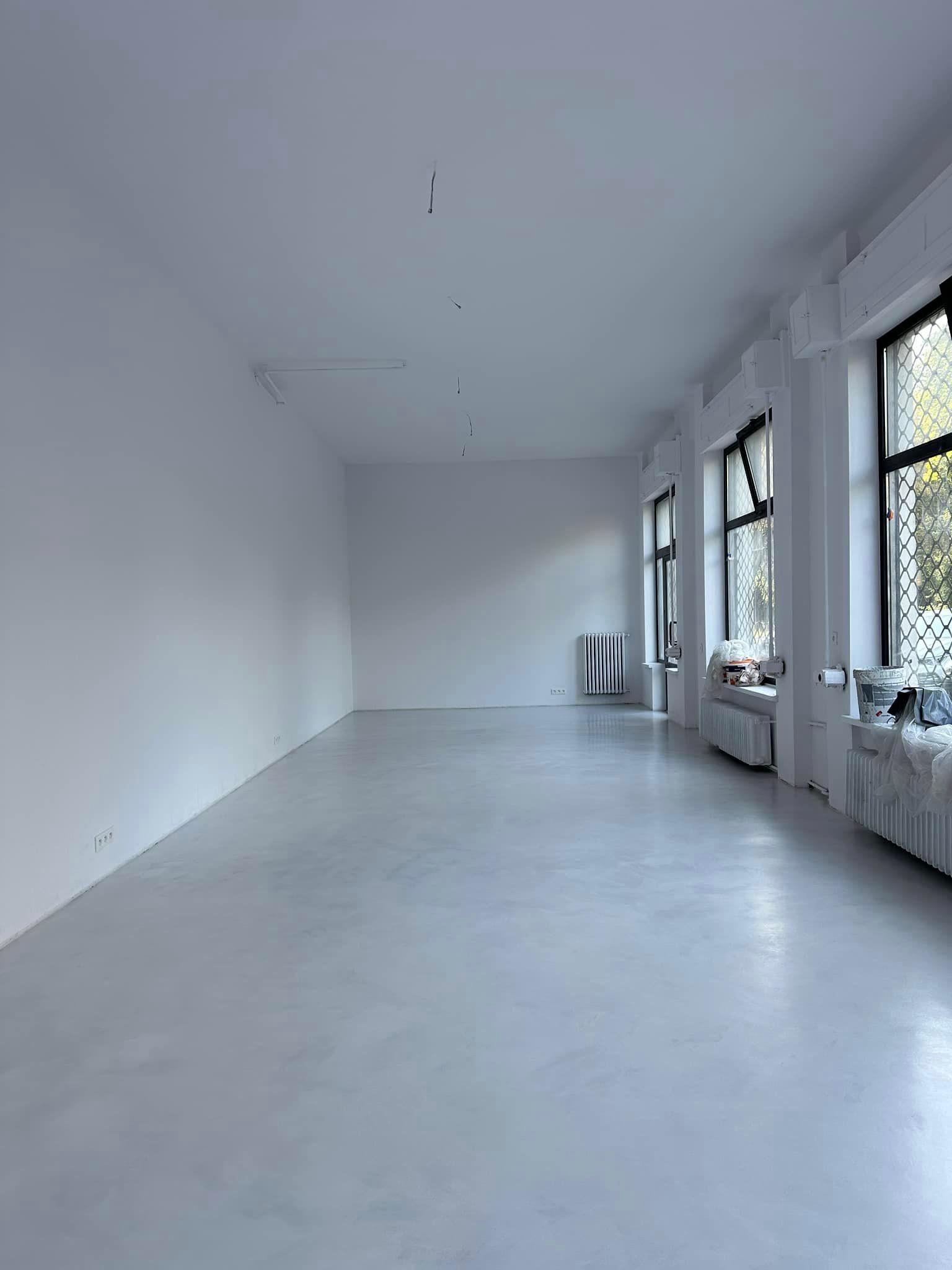 Jasnoszara, gładka posadzka w pustym pomieszczeniu z białymi ścianami i trzema oknami z kratami. Widoczne grzejniki pod oknami. Minimalistyczny wystrój.