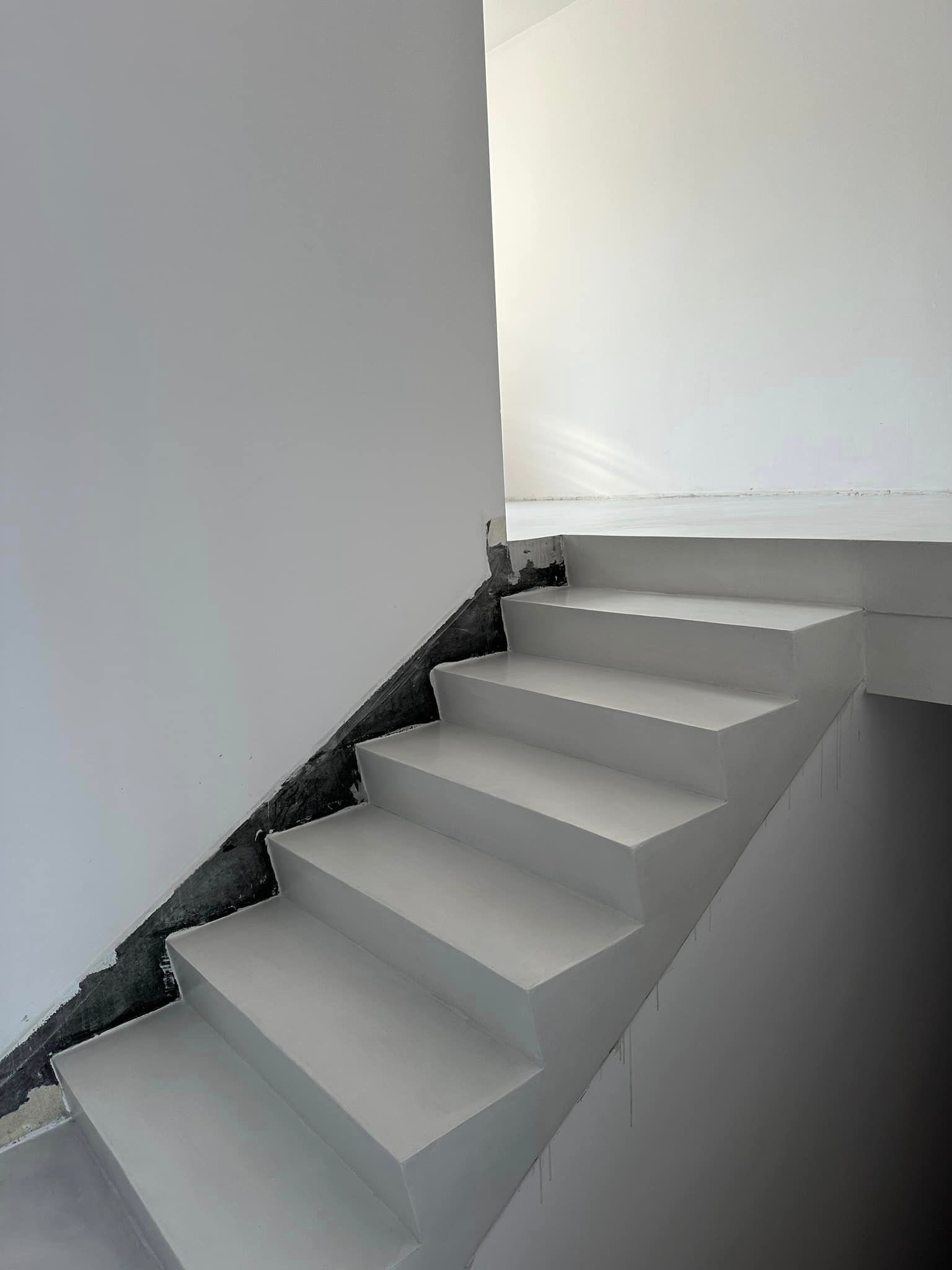 Schody wewnętrzne pokryte jasnoszarą posadzką żywiczną, widoczne niedoskonałości wykończenia przy ścianie, ślady spływającej farby na ścianie bocznej.