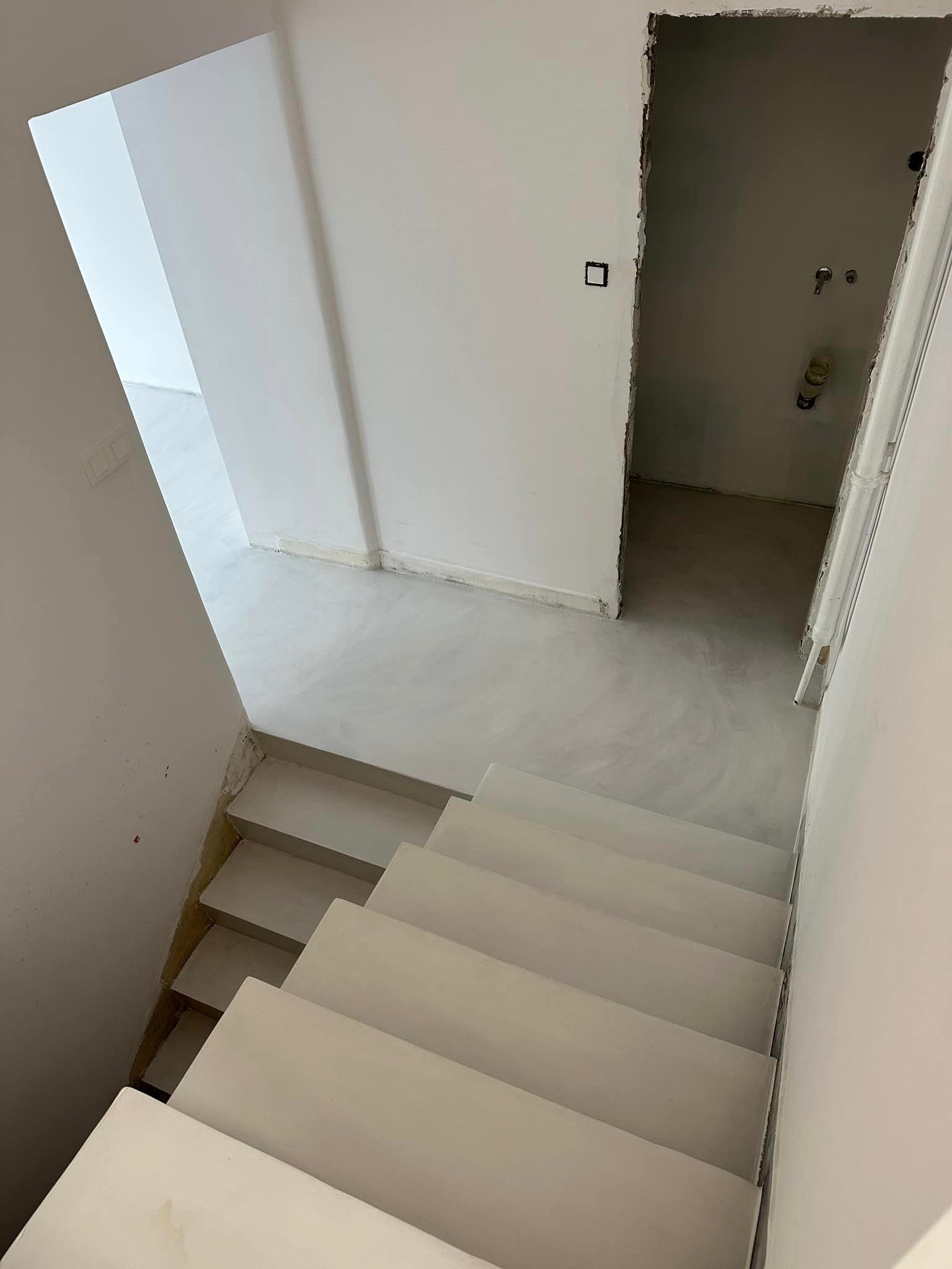 Widok z góry na białe schody w surowym stanie, z szarą podłogą i ścianami w tle, w trakcie remontu lub budowy.