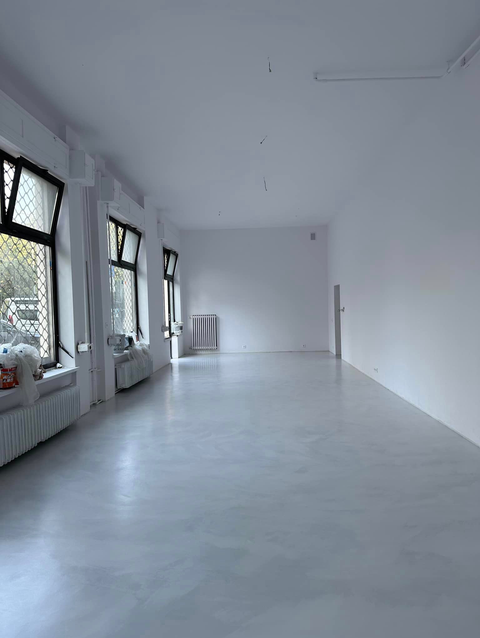 Duże, jasne pomieszczenie z szarą, błyszczącą podłogą, prawdopodobnie w trakcie wykańczania, z widocznymi grzejnikami pod oknami i workami budowlanymi w rogu.