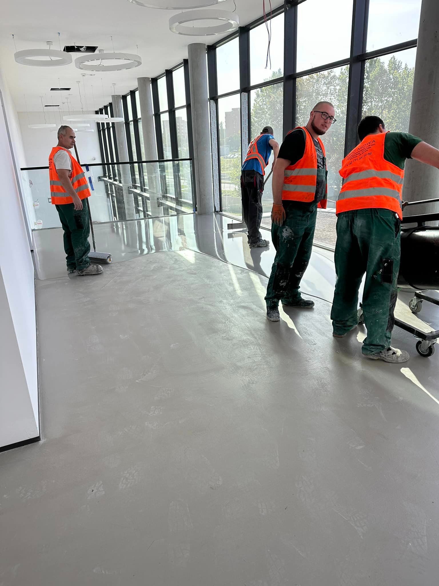 Praca wre: ekipa w pomarańczowych kamizelkach aplikuje szarą posadzkę w przestronnym, jasnym wnętrzu z kolumnami i dużymi oknami. Widoczne narzędzia i ślady butów.