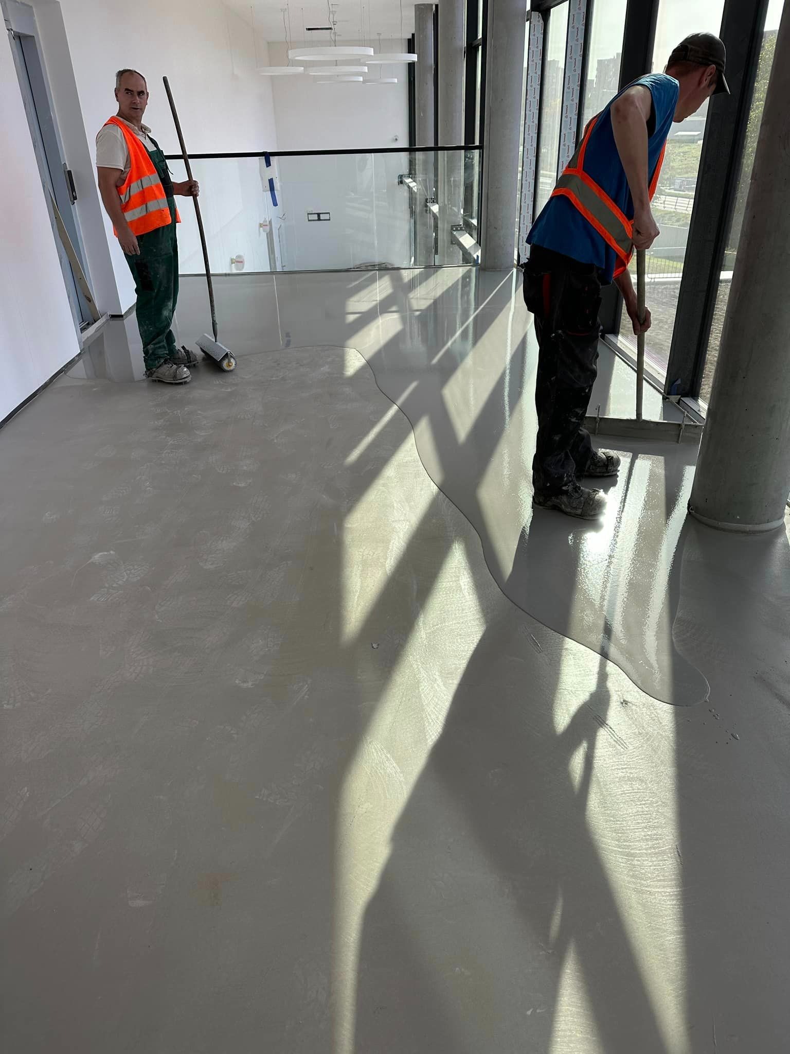 Aplikacja szarej, błyszczącej posadzki w jasnym wnętrzu. Dwóch pracowników w odblaskowych kamizelkach rozprowadza materiał na podłodze wzdłuż okien.