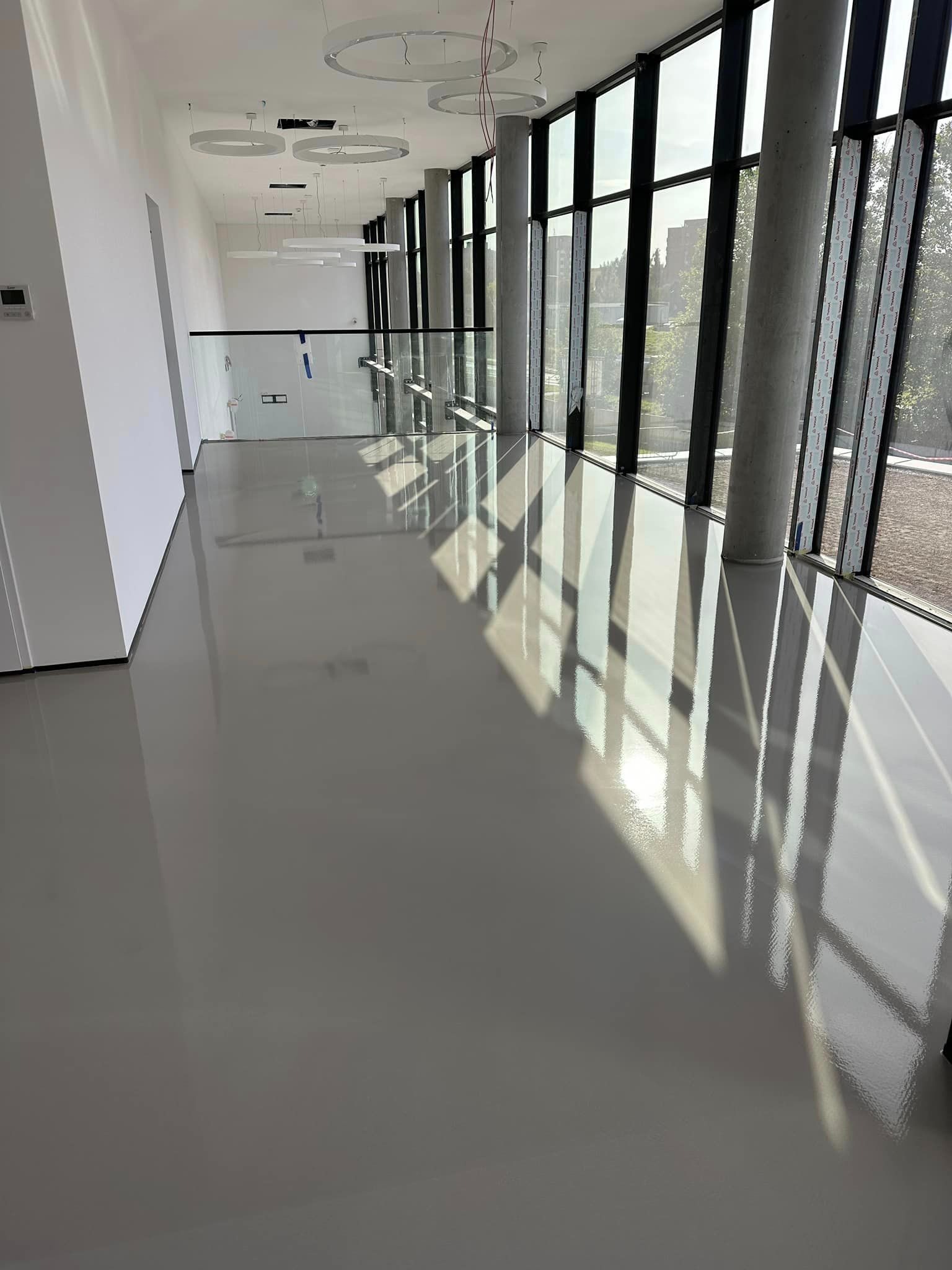 Jasnoszara, błyszcząca posadzka w przestronnym wnętrzu z dużymi oknami. Widoczne odbicia światła na gładkiej powierzchni. Elementy szklanej balustrady w tle.